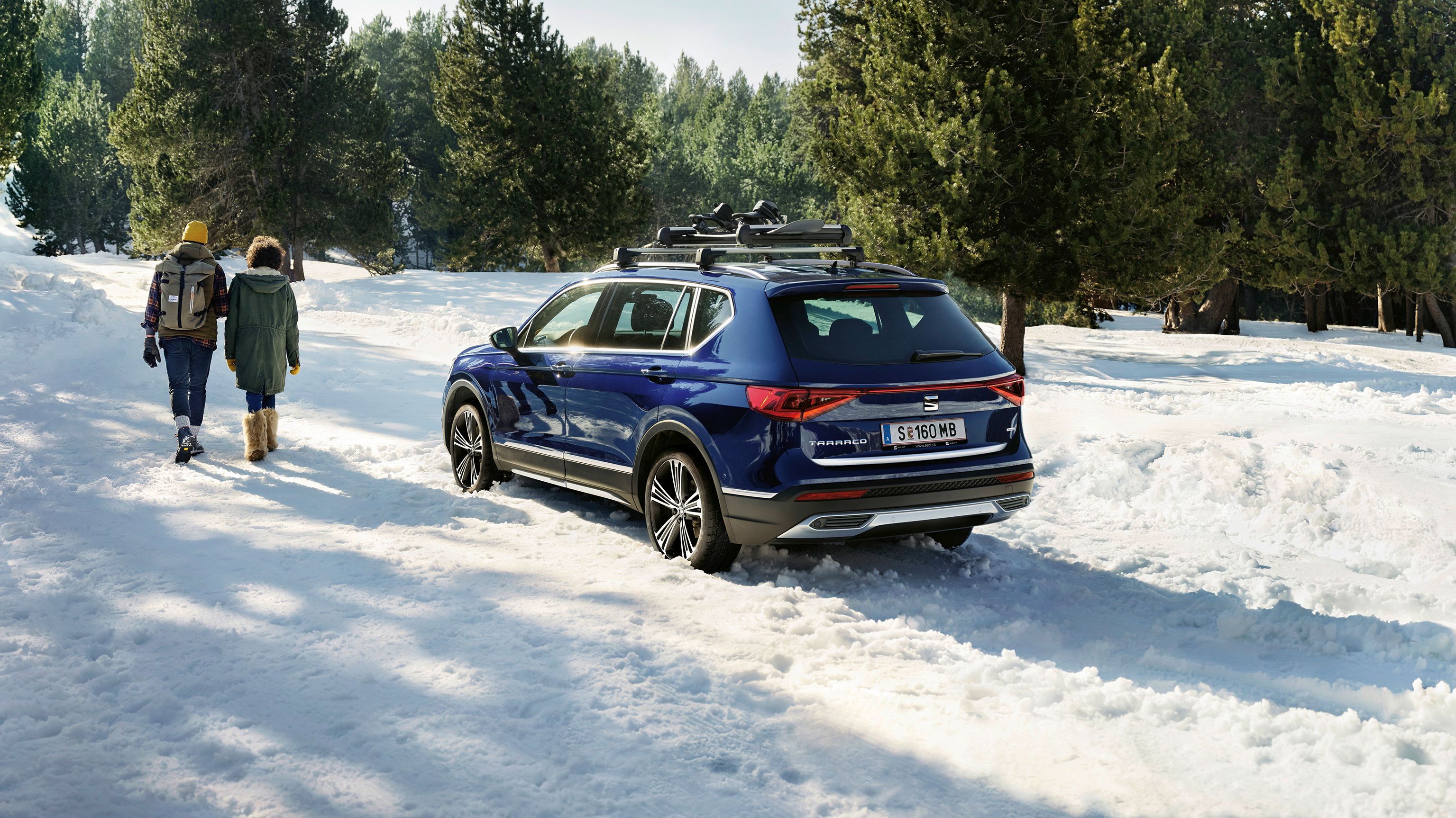 Ein blauer SEAT Tarraco mit Dachträger parkt an einem verschneiten Weg. Ein Mann und eine Frau wandern vom Fahrzeug fort.