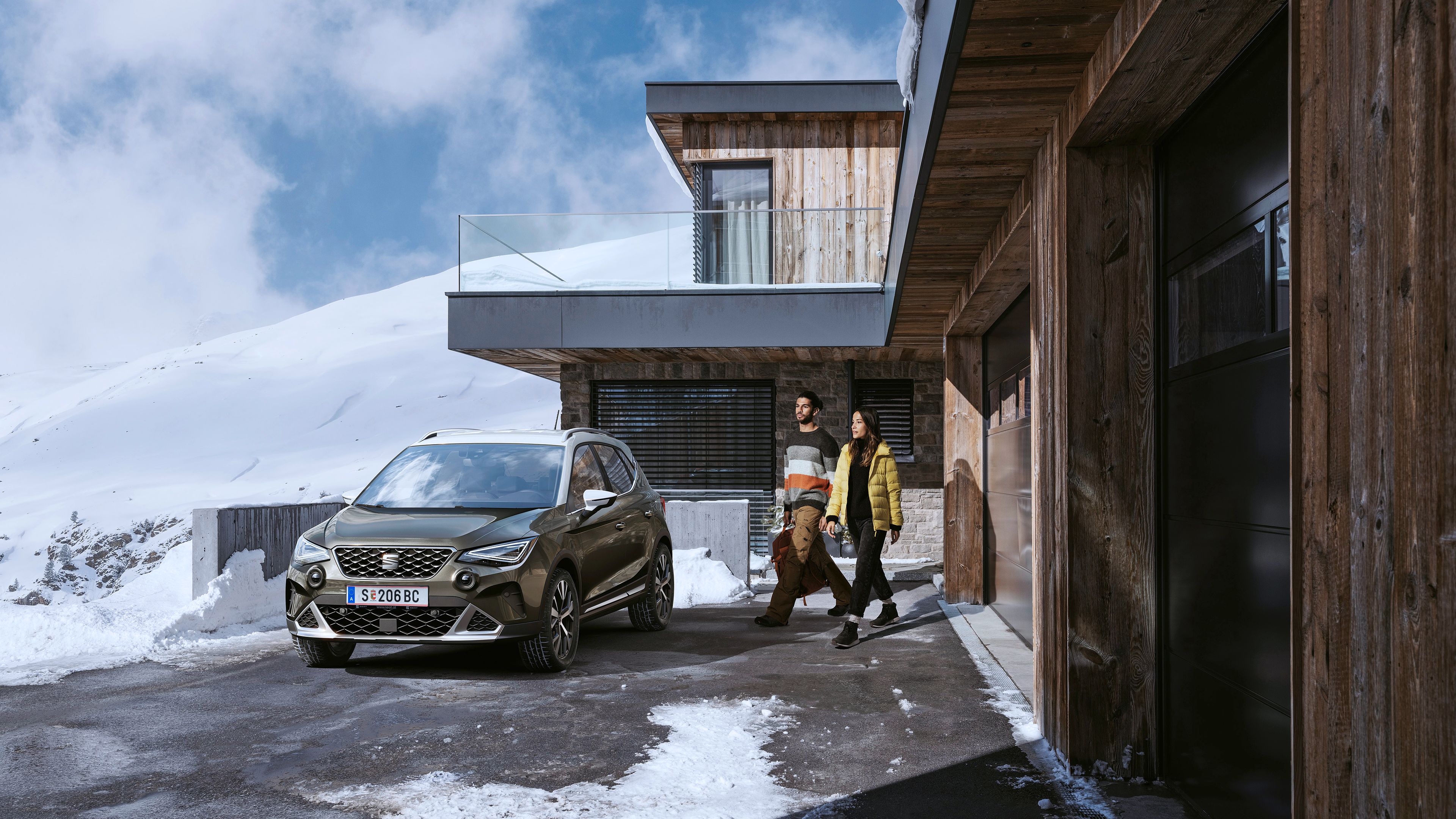 Ein Mann und eine Frau auf dem Weg zu ihrem SEAT Arona, der vor einem Haus in den winterlichen Bergen geparkt ist.