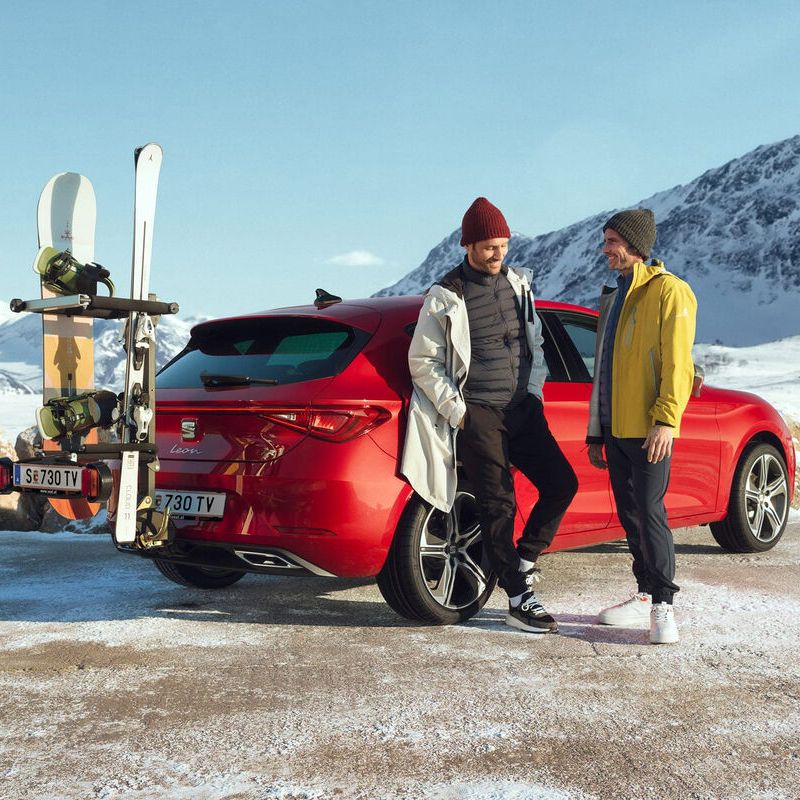 Zwei Männer vor einem roten SEAT Leon mit Anhängerkupplung und Skiträger in einer winterlichen Berglandschaft.