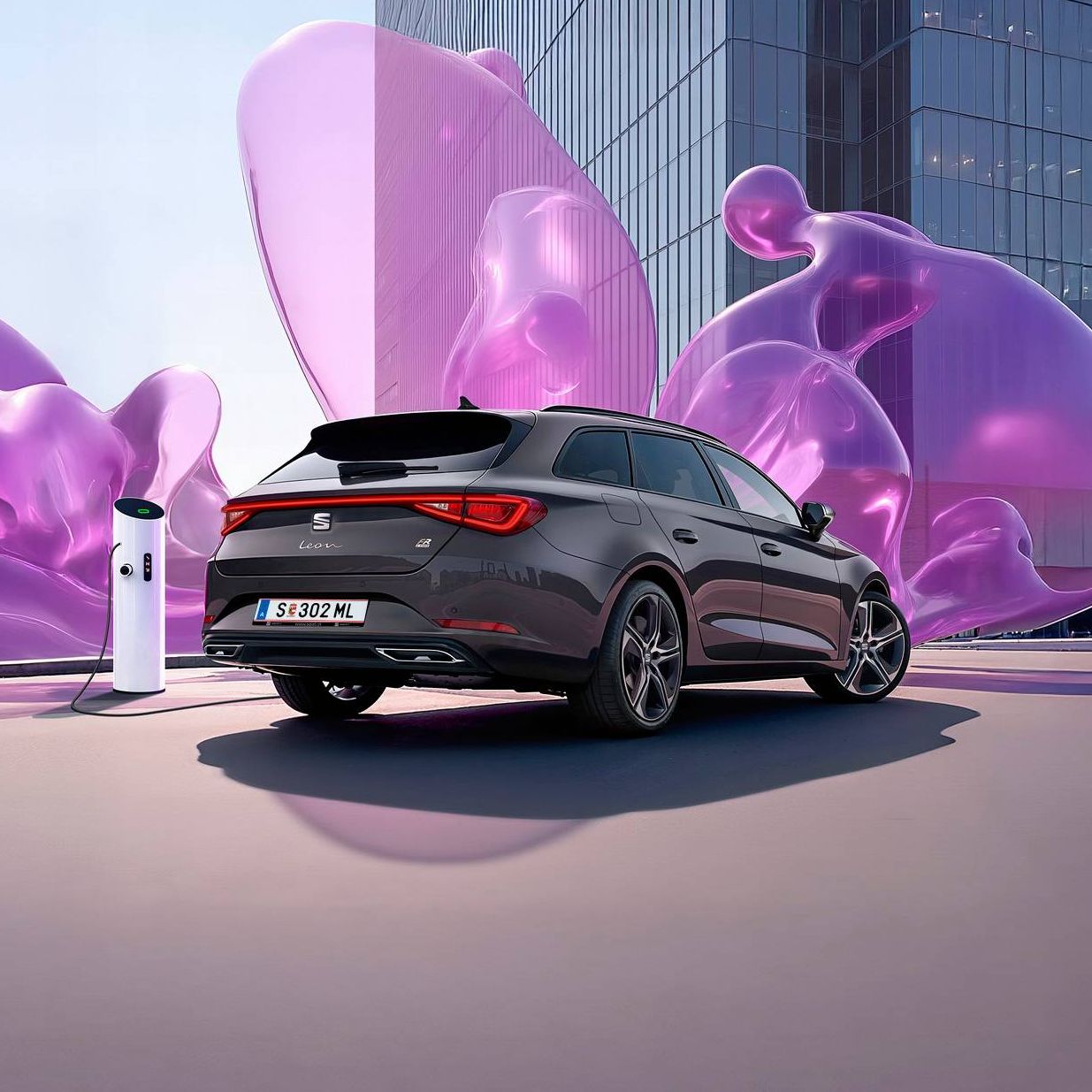 Schräge Heckansicht eines dunkelgrauen SEAT Leon SP Kombi beim Laden an einer Ladesäule vor einem modernen Gebäude mit Glasfronten.