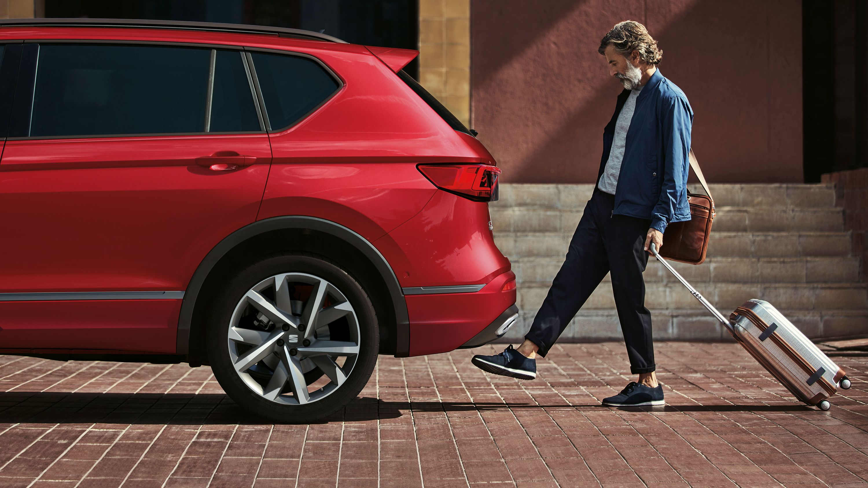 Ein Mann mit Rollkoffer betätigt mit dem Fuß die Kick-to-Open Funktion zum Öffnen des Kofferraums bei einem SEAT Tarraco. 