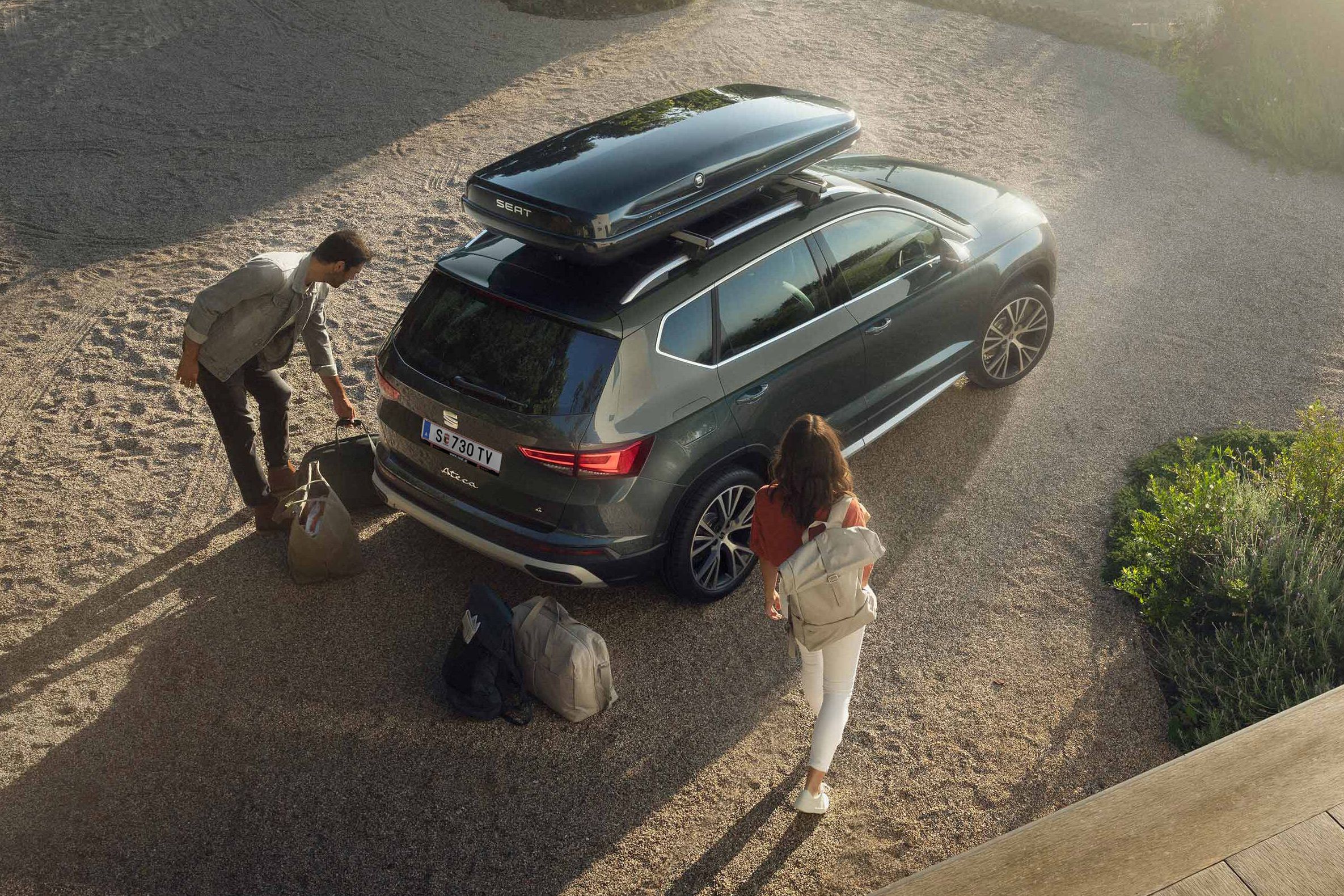 Ein SEAT Ateca mit einer schwarzen Dachbox. Ein Mann und eine Frau mit Reisetaschen dahinter bereiten die Abreise vor.