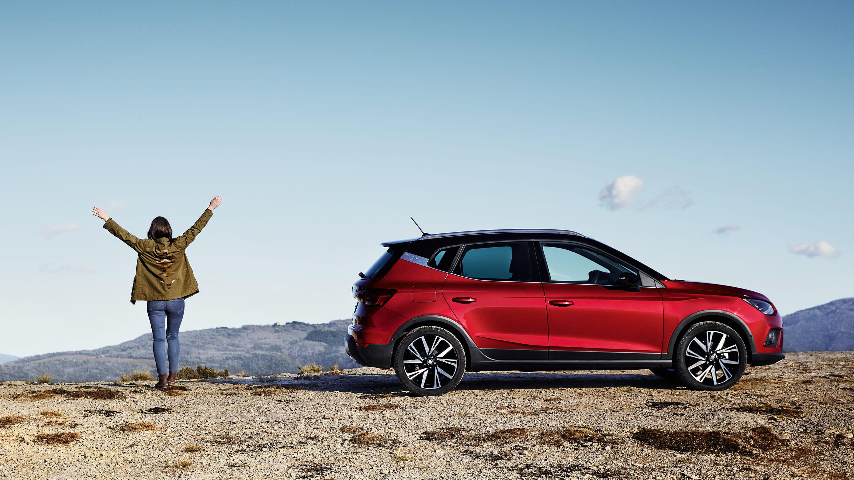 Eine Frau streckt die Arme hoch und blickt über die Landschaft. Neben ihr ein roter SEAT Arona in Seitenansicht.
