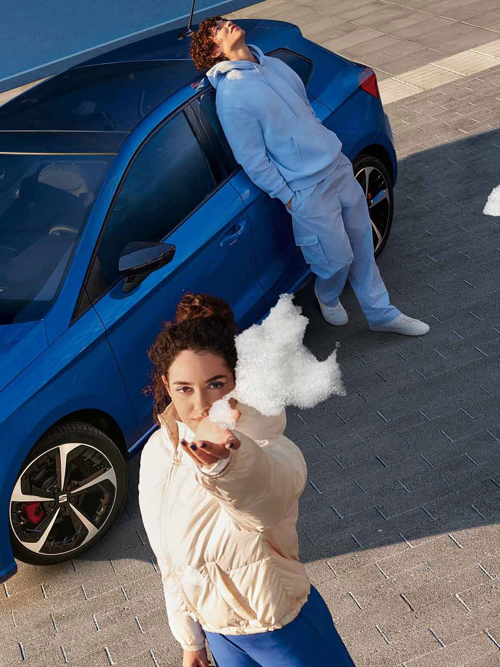 Ein blauer SEAT Ibiza aus der Vogelperspektive: Drei Personen, alle in blauer Kleidung, posieren am Auto – zwei lehnen an der Motorhaube, eine an der Seitentür. Eine der Personen hält Schaum in die Kamera.
