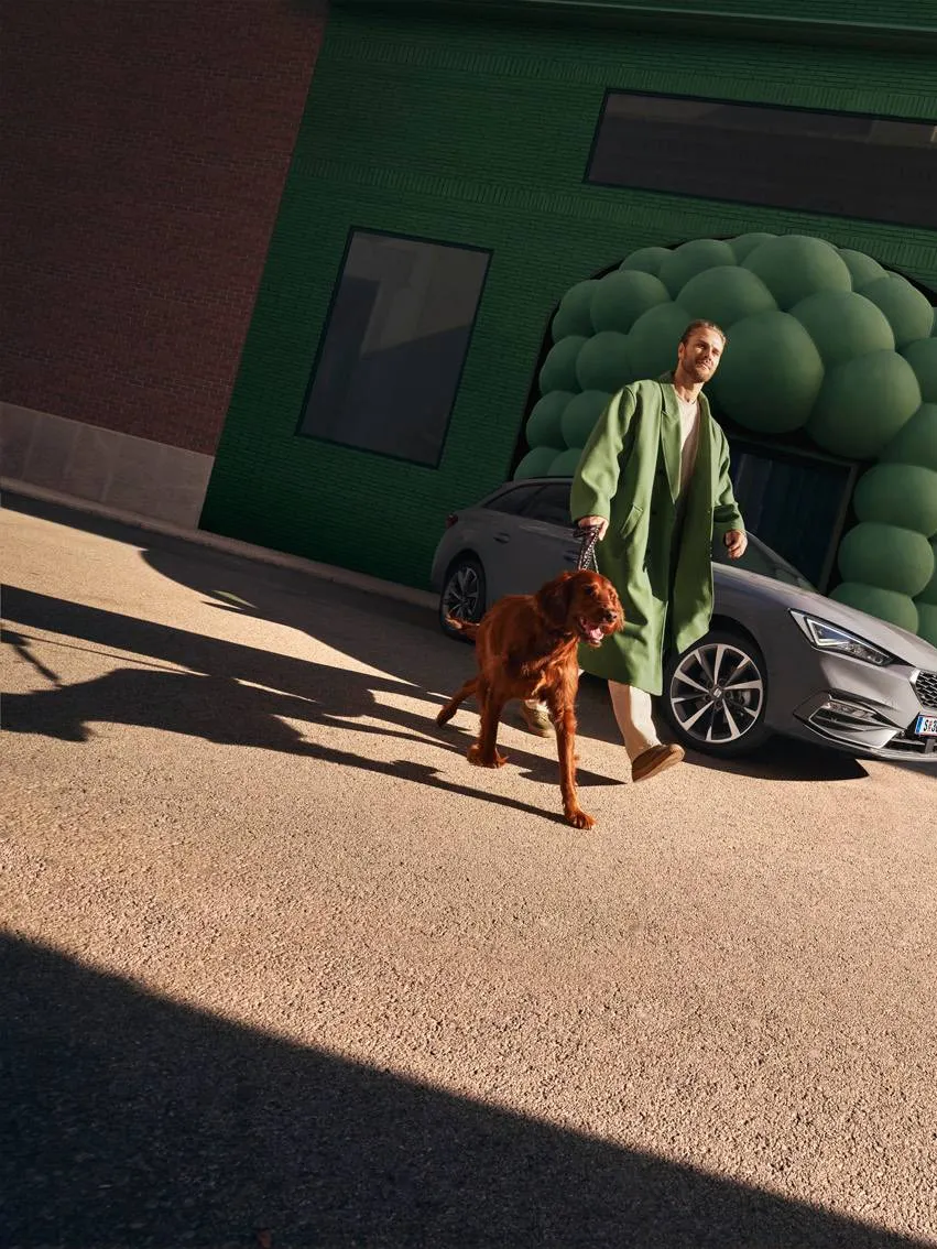 Ein Mann geht mit seinem Hund spazieren. Im Hintergrund ist sein SEAT Leon Kombi zu sehen.