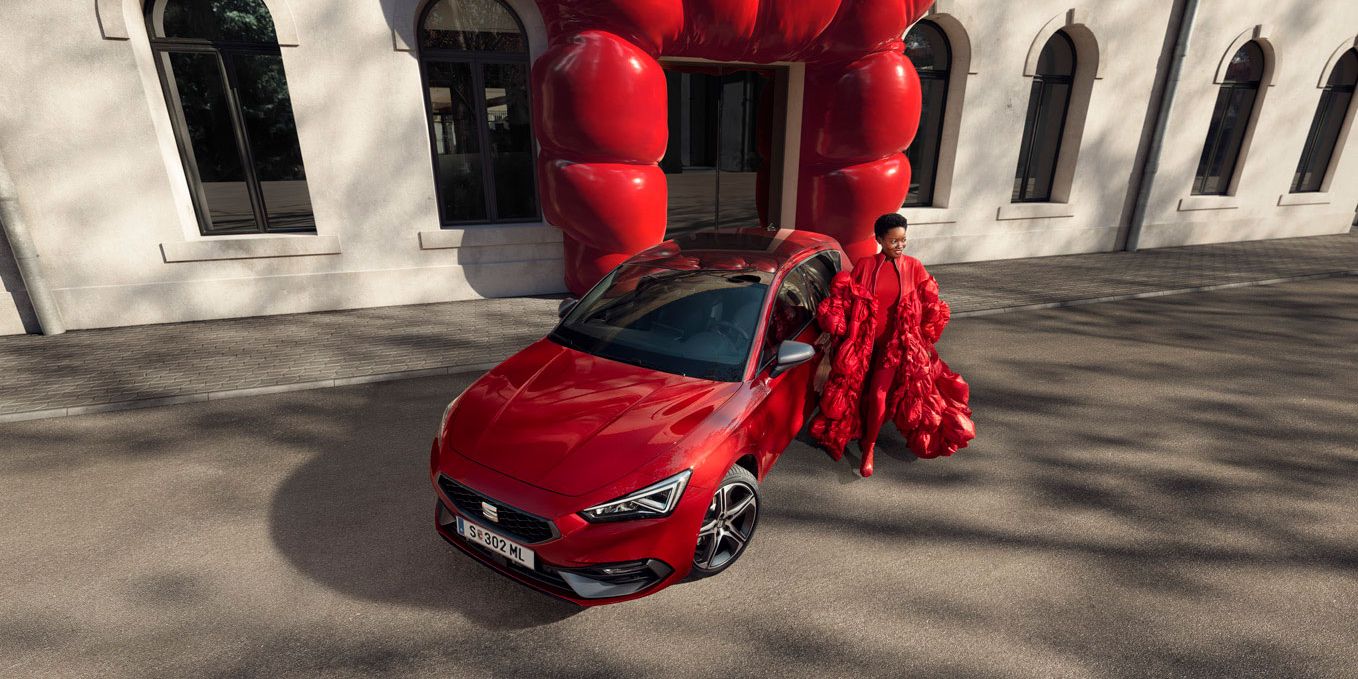 Ein roter SEAT Leon 5-Türer steht vor einem Gebäude, eine Frau in einem roten Kleid steht rechts neben dem Auto.