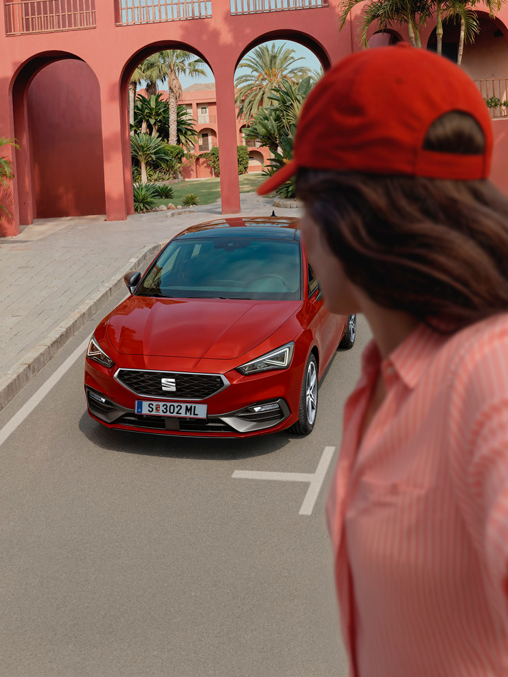 SEAT Leon vor Gebäude und Mädchen steht verkehrt davor.