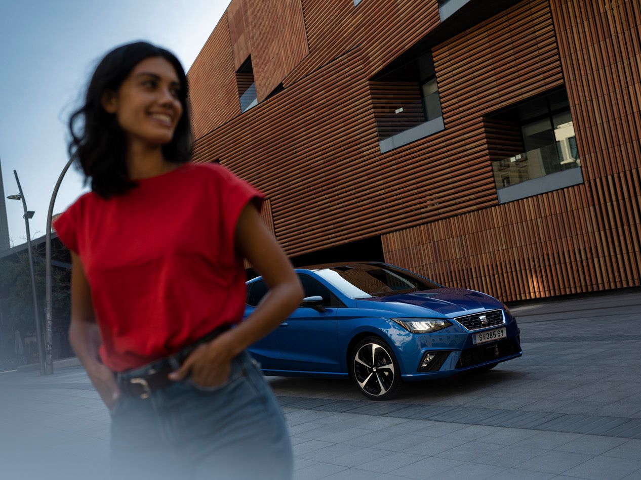 Eine Frau steht vor einem blauen SEAT Ibiza, wobei die Frau verschwommen ist und das SEAT Ibiza Modell im Fokus steht 