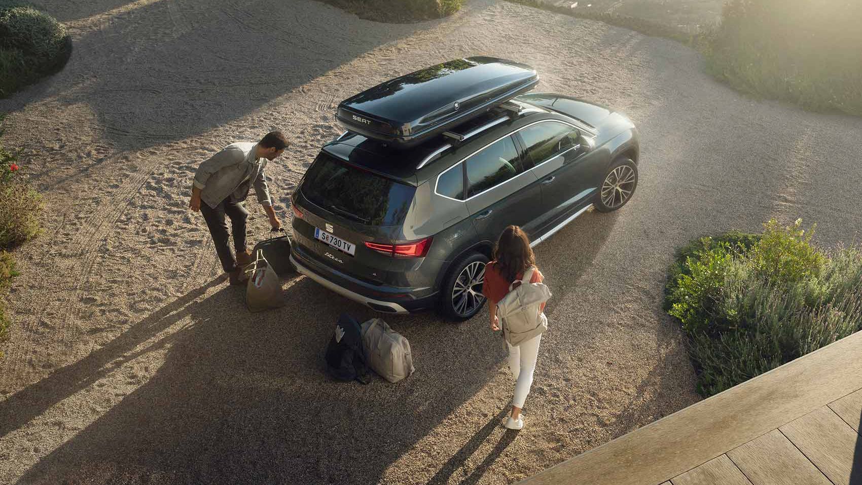 Ein SEAT Ateca mit einer schwarzen Dachbox. Ein Mann und eine Frau mit Reisetaschen dahinter bereiten die Abreise vor.