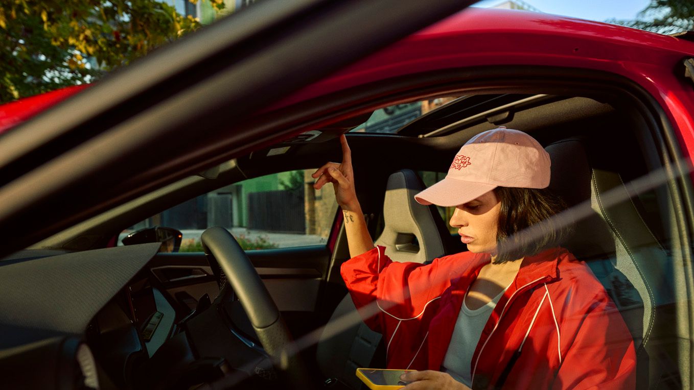 Eine Person in roter Jacke und rosa Mütze sitzt in einem roten SEAT und hält ein gelbes Smartphone. Sie zeigt mit einer Hand nach oben.
