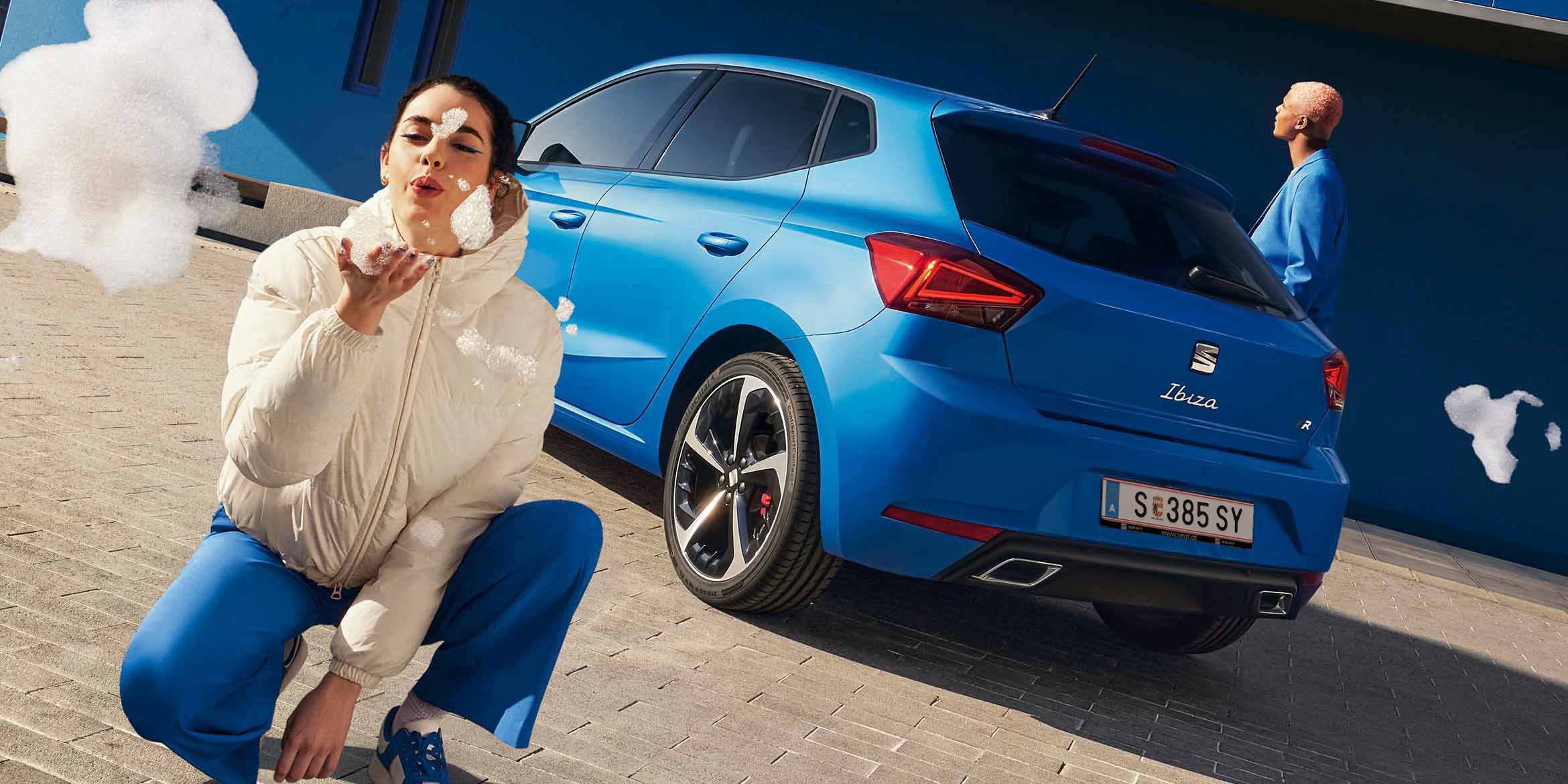 Ein blauer SEAT Ibiza steht mit dem Heck zu einem blauen Gebäude. Ein Mann im blauen Anzug geht daran vorbei, während eine Frau direkt vor der Kamera kniet und Seifenblasen in Richtung Linse pustet.