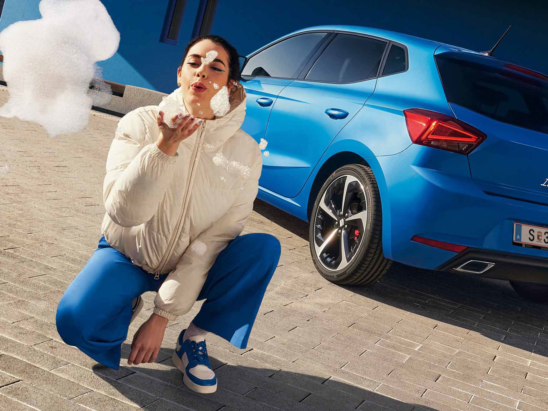 Ein blauer SEAT Ibiza steht mit dem Heck zu einem blauen Gebäude. Ein Mann im blauen Anzug geht daran vorbei, während eine Frau direkt vor der Kamera kniet und Seifenblasen in Richtung Linse pustet.