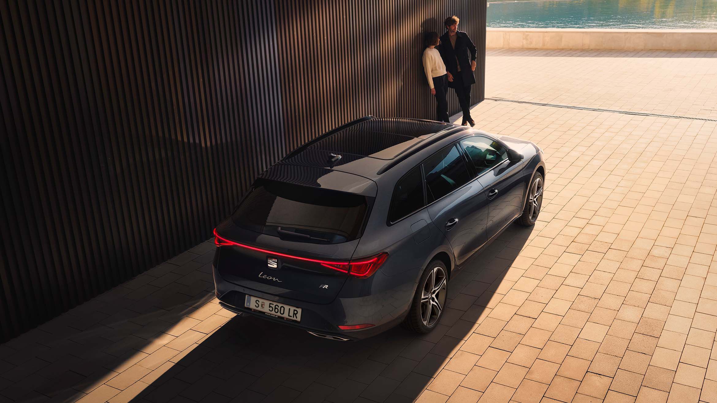 Heckansicht eines SEAT Leon SP Kombi inder Farbe Magnetic Tech. Links vom Fahrzeug lehnen ein Mann und eine Frau an einer Wand.