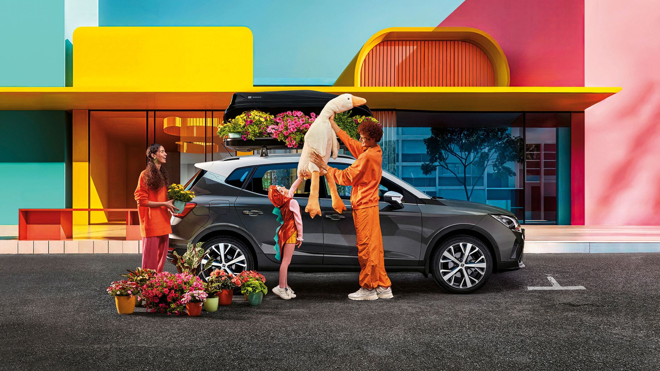 Eine Familie mit Blumen und einem Stofftier befindet sich vor einem SEAT Arona mit Dachbox.