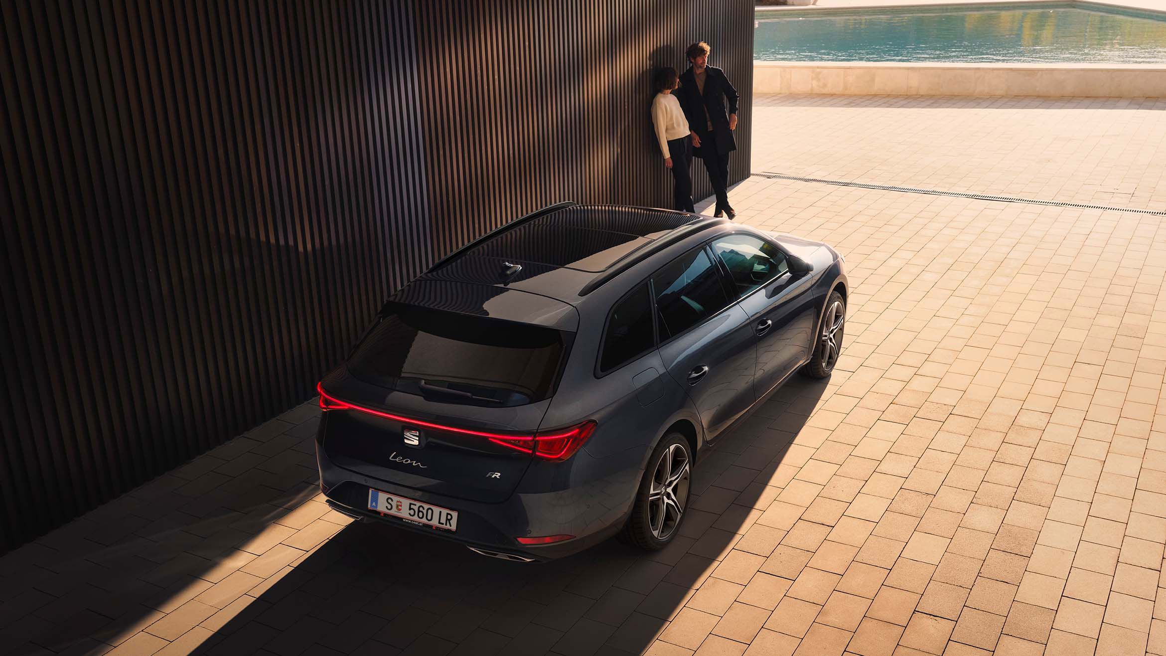 Heckansicht eines SEAT Leon SP Kombi inder Farbe Magnetic Tech. Links vom Fahrzeug lehnen ein Mann und eine Frau an einer Wand.