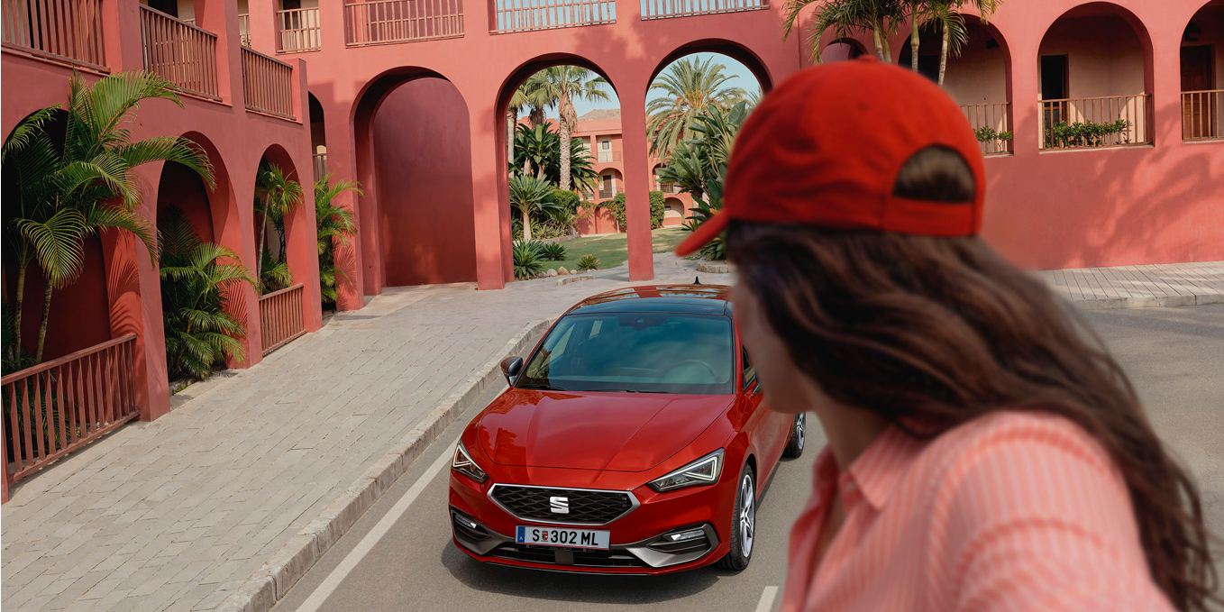 [AI-modified] – SEAT Leon vor Gebäude und Mädchen steht verkehrt davor.