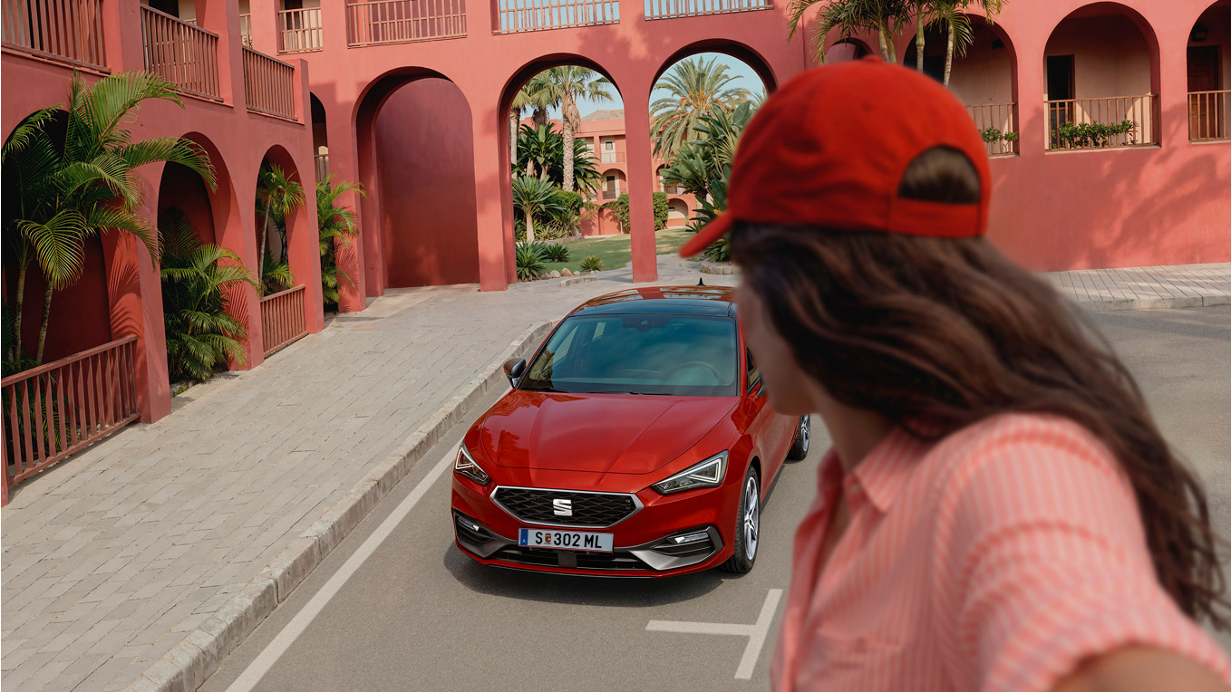 [AI-modified] – SEAT Leon vor Gebäude und Mädchen steht verkehrt davor.