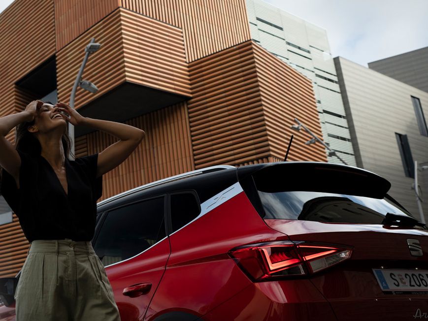Eine Frau steht vor einem roten SEAT Arona mit schwarzem Dach, der aus der Heckperspektive zu sehen ist