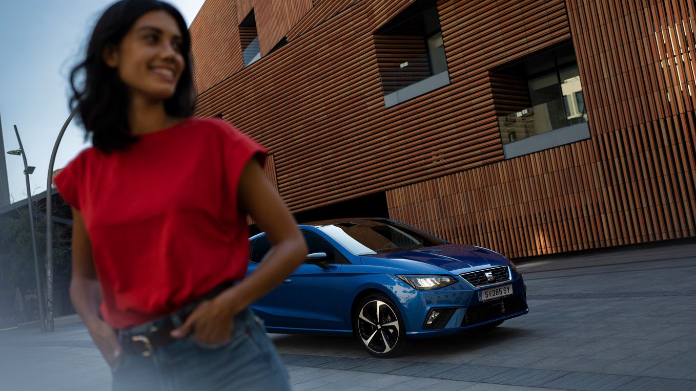 Eine Frau steht vor einem blauen SEAT Ibiza, wobei die Frau verschwommen ist und das SEAT Ibiza Modell im Fokus steht 