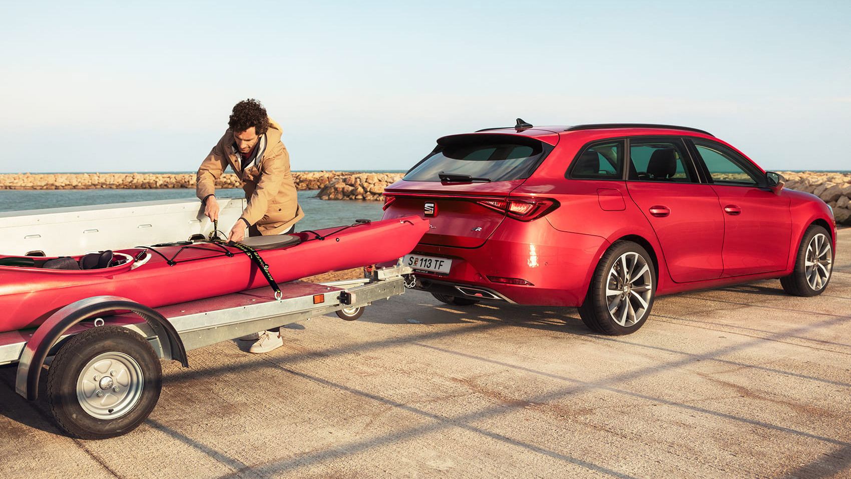 Ein roter SEAT Leon SP Kombi mit Anhänger an der Küste. Ein Mann montiert ein Kanu auf dem Anhänger.