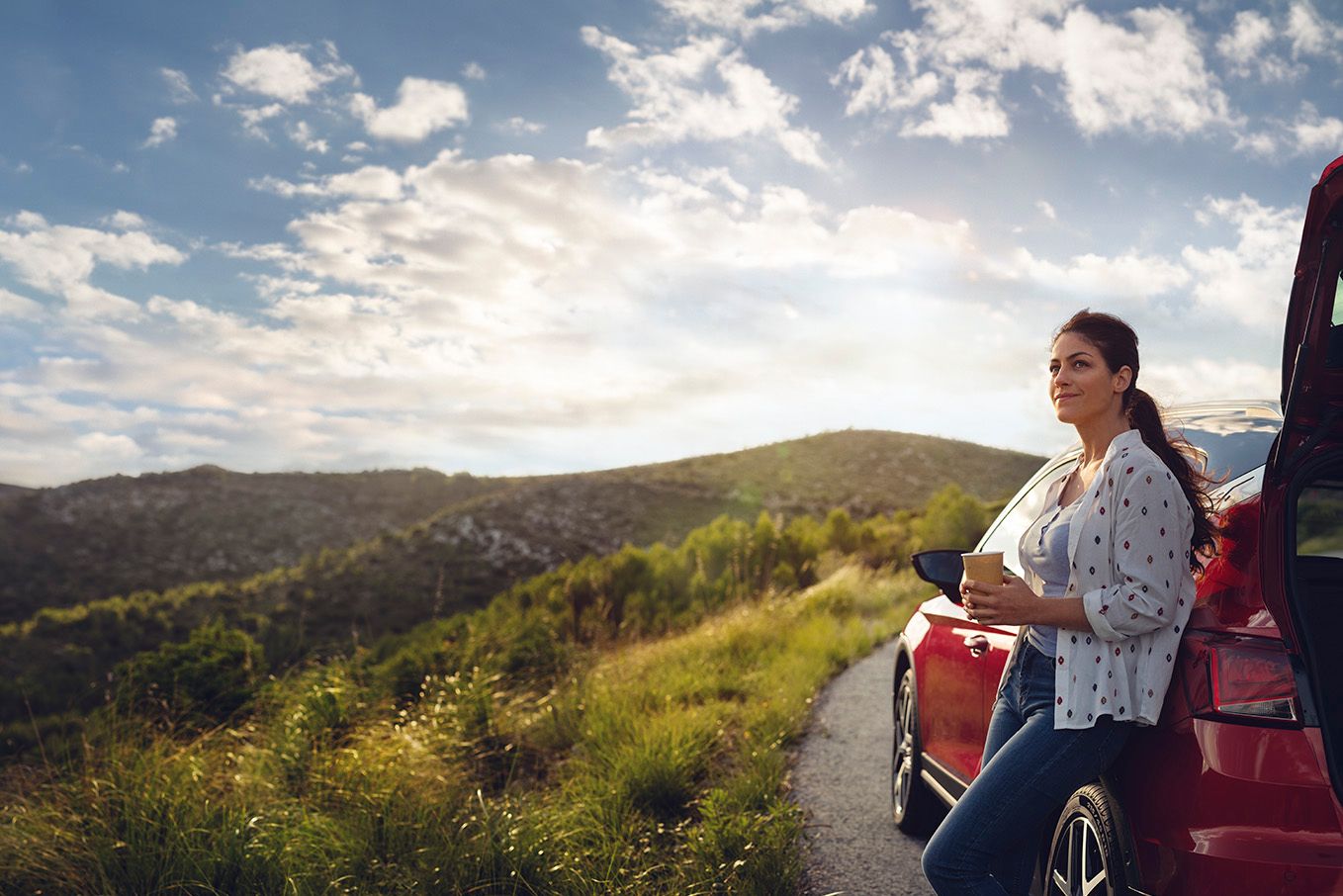Eine Frau lehnt an einem roten SEAT Ibiza, hält einen Becher in der Hand und blickt auf die umliegende Berglandschaft entlang einer Landstraße.