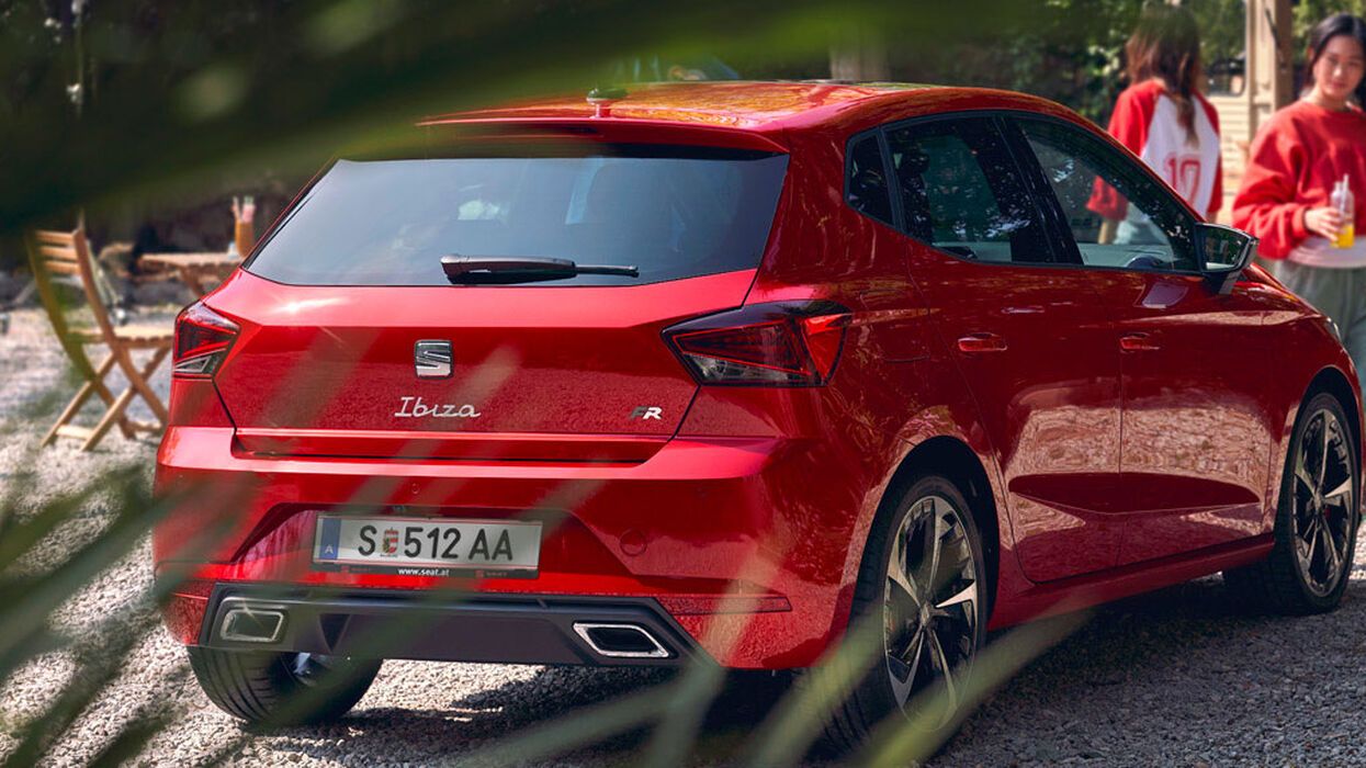 Roter SEAT Ibiza steht auf einen Schotterplatz und im Hintergrund sind Frauen mit einem Getränk in der Hand. 