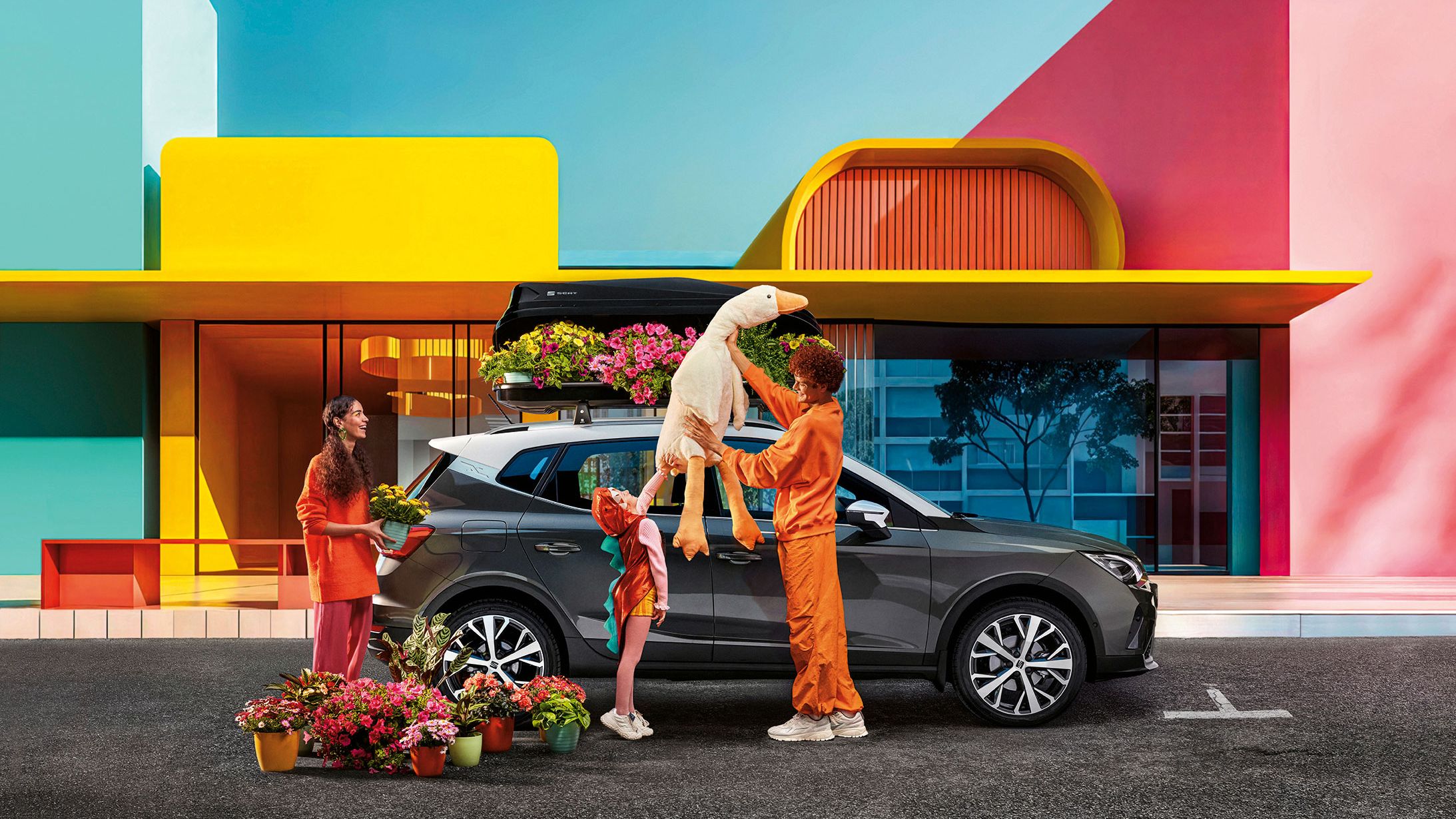 Eine Familie mit Blumen und einem Stofftier befindet sich vor einem SEAT Arona mit Dachbox.