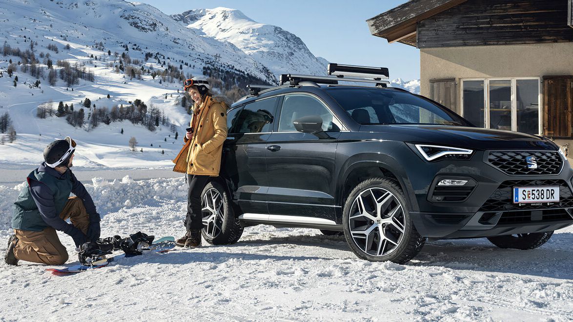 Ein SEAT Ateca in winterlicher Landschaft. Eine Frau lehnt am Fahrzeug und ein Mann bereitet ein Snowboard vor.