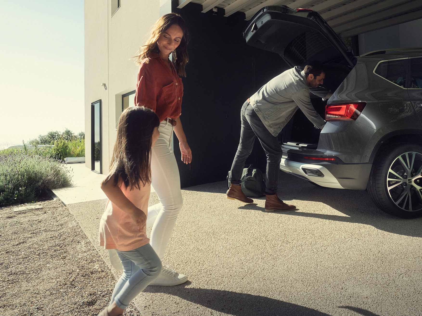Ein Mann belädt den Kofferraum eines grauen SEAT Ateca. Die Mutter und ihre Tochter freuen sich schon auf die Abfahrt. 