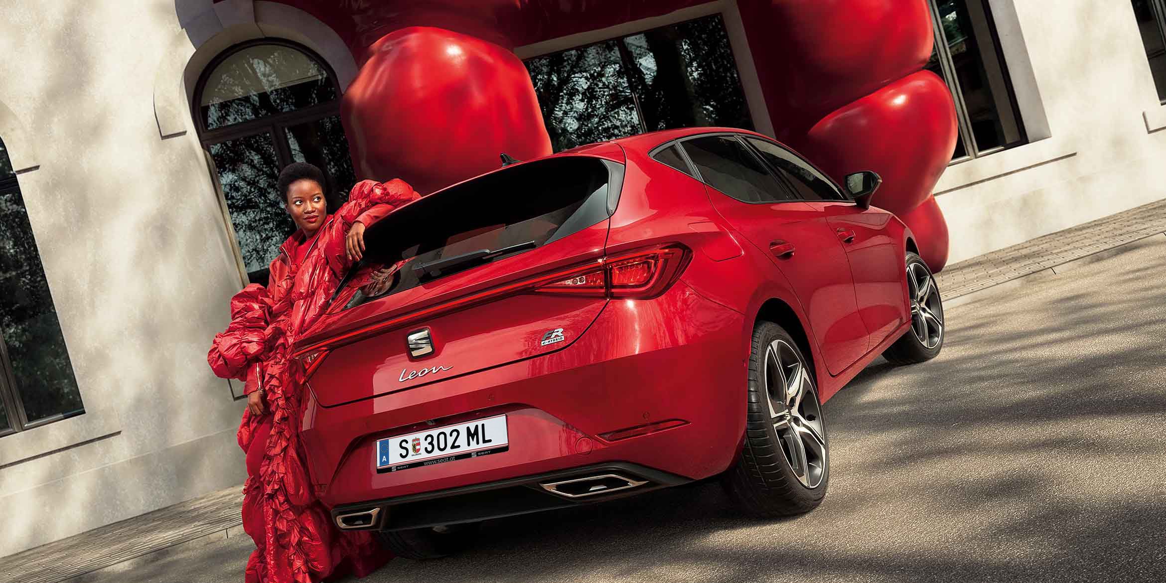 Eine stilvoll in Rot gekleidete Frau lehnt lässig an einem roten SEAT Leon FR e-HYBRID mit modernen LED-Rückleuchten