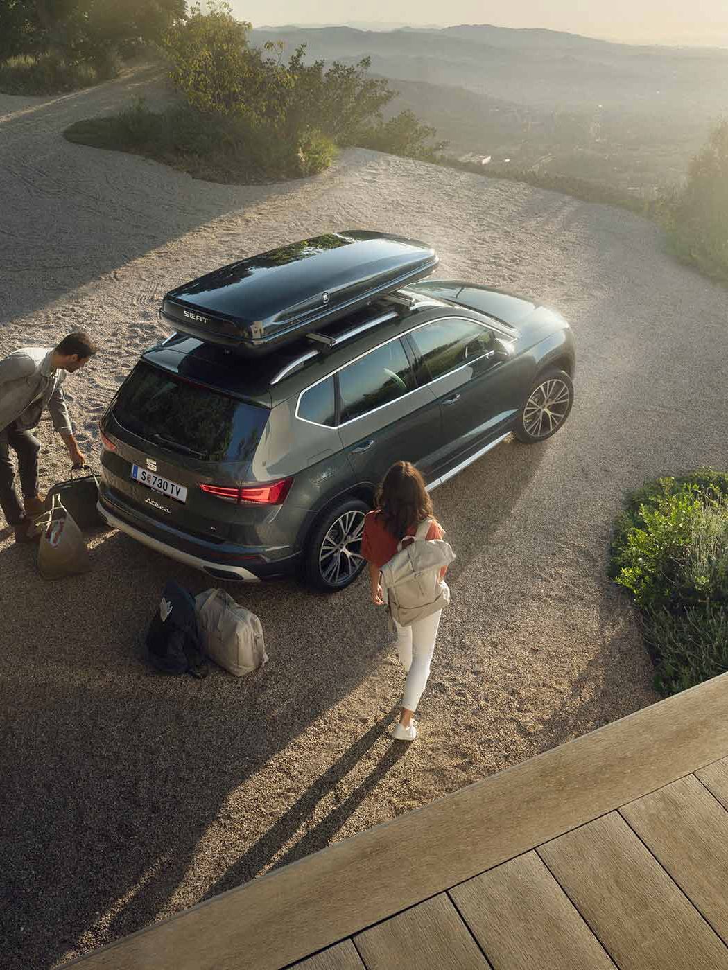 Ein SEAT Ateca mit einer schwarzen Dachbox. Ein Mann und eine Frau mit Reisetaschen dahinter bereiten die Abreise vor.